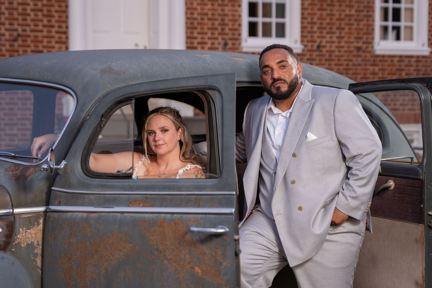 A couple poses with a vintage rusty pickup truck, the bride in the driver's seat while her partner stands outside.