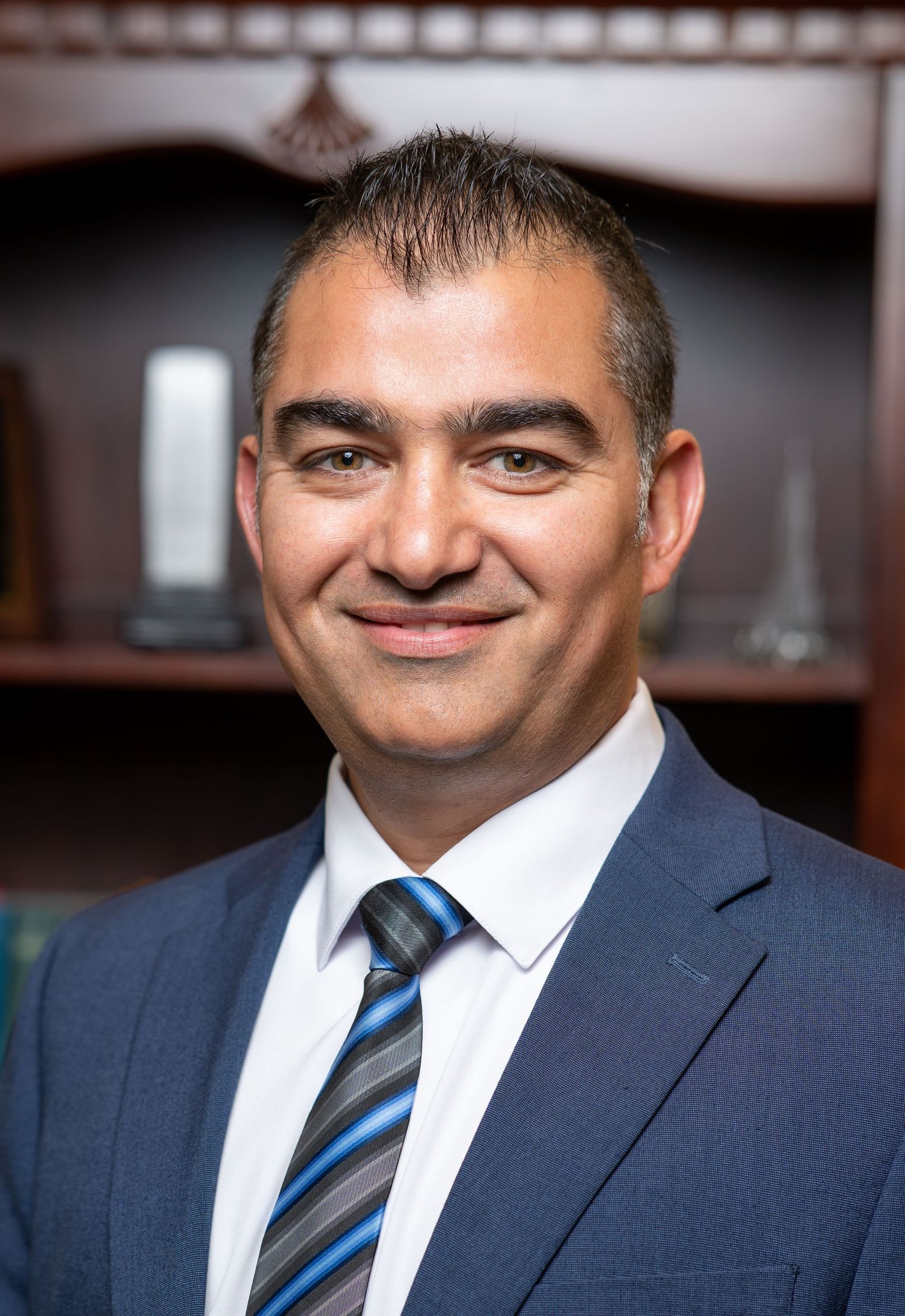 Professional headshot of a smiling businessman in a navy suit and striped tie against an office bookshelf background.