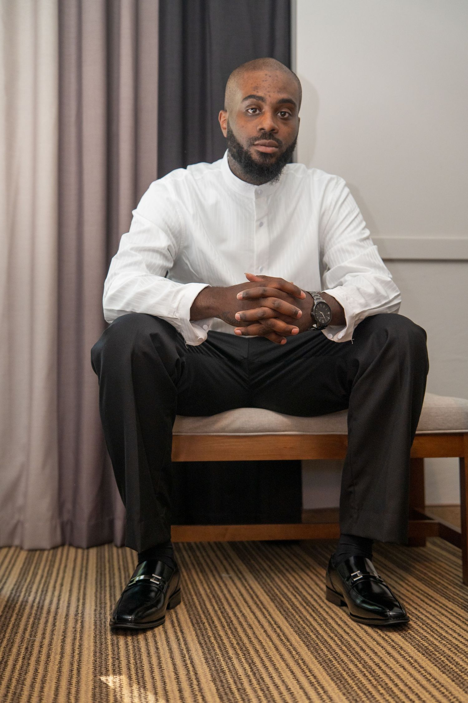 A person in a white sweater and black pants sits on a wooden bench against a neutral wall with striped carpeting.