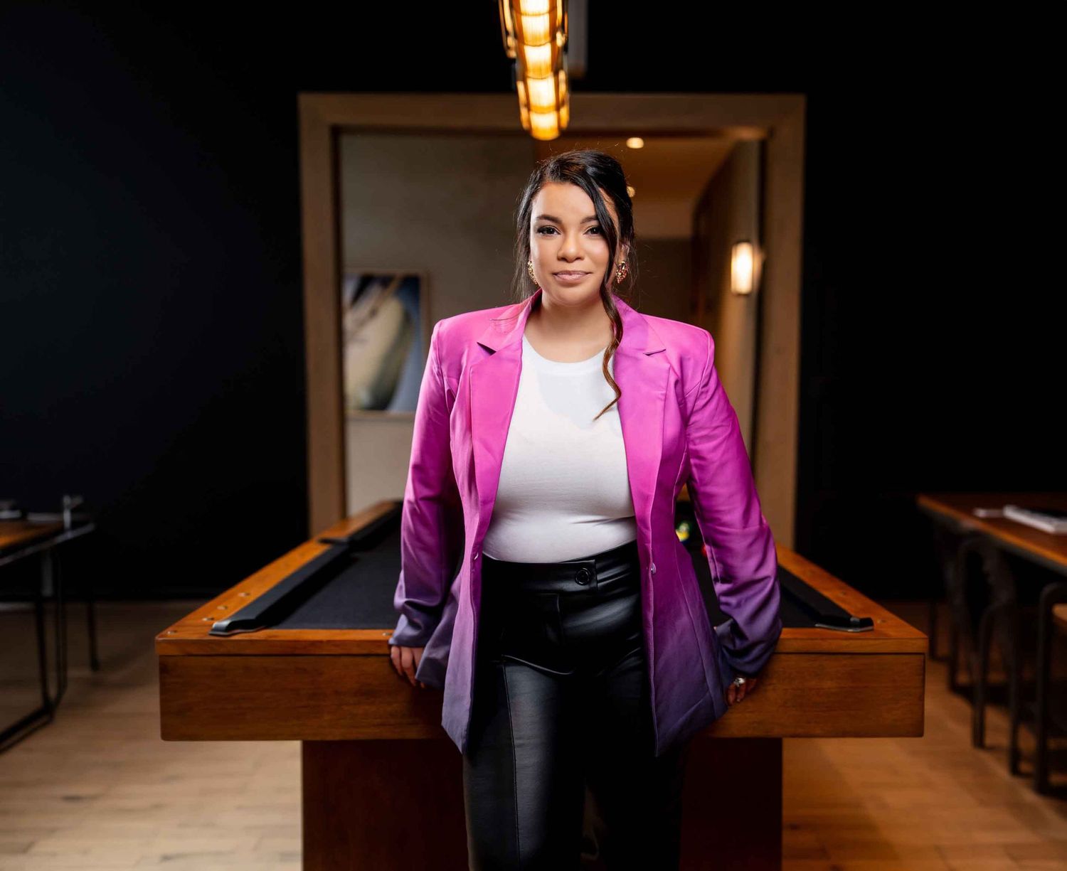 Professional wearing a vibrant magenta blazer and white top poses in a warmly lit office space with wooden furniture.