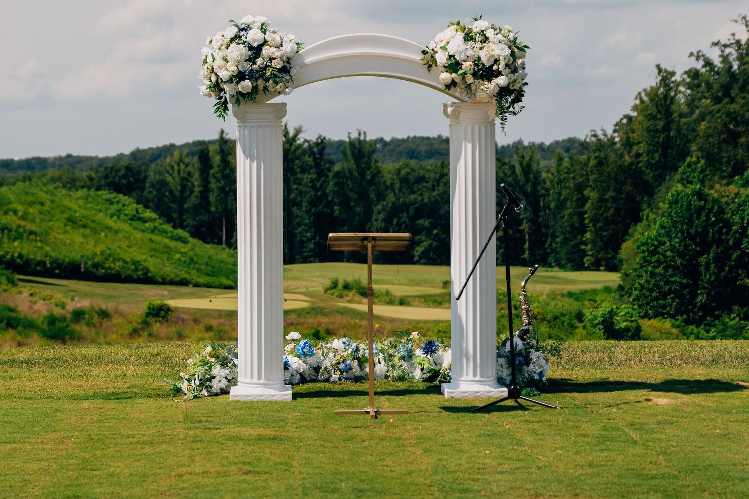 White wedding arch decorated with floral arrangements stands against a scenic backdrop of rolling green hills and trees.
