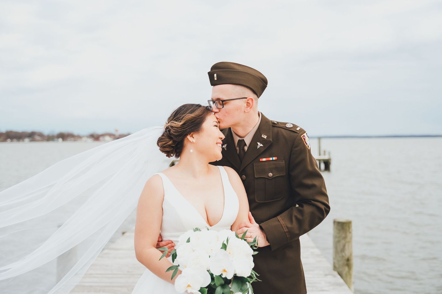 A couple in wedding attire embraces on a dock overlooking a serene waterfront on their special day.