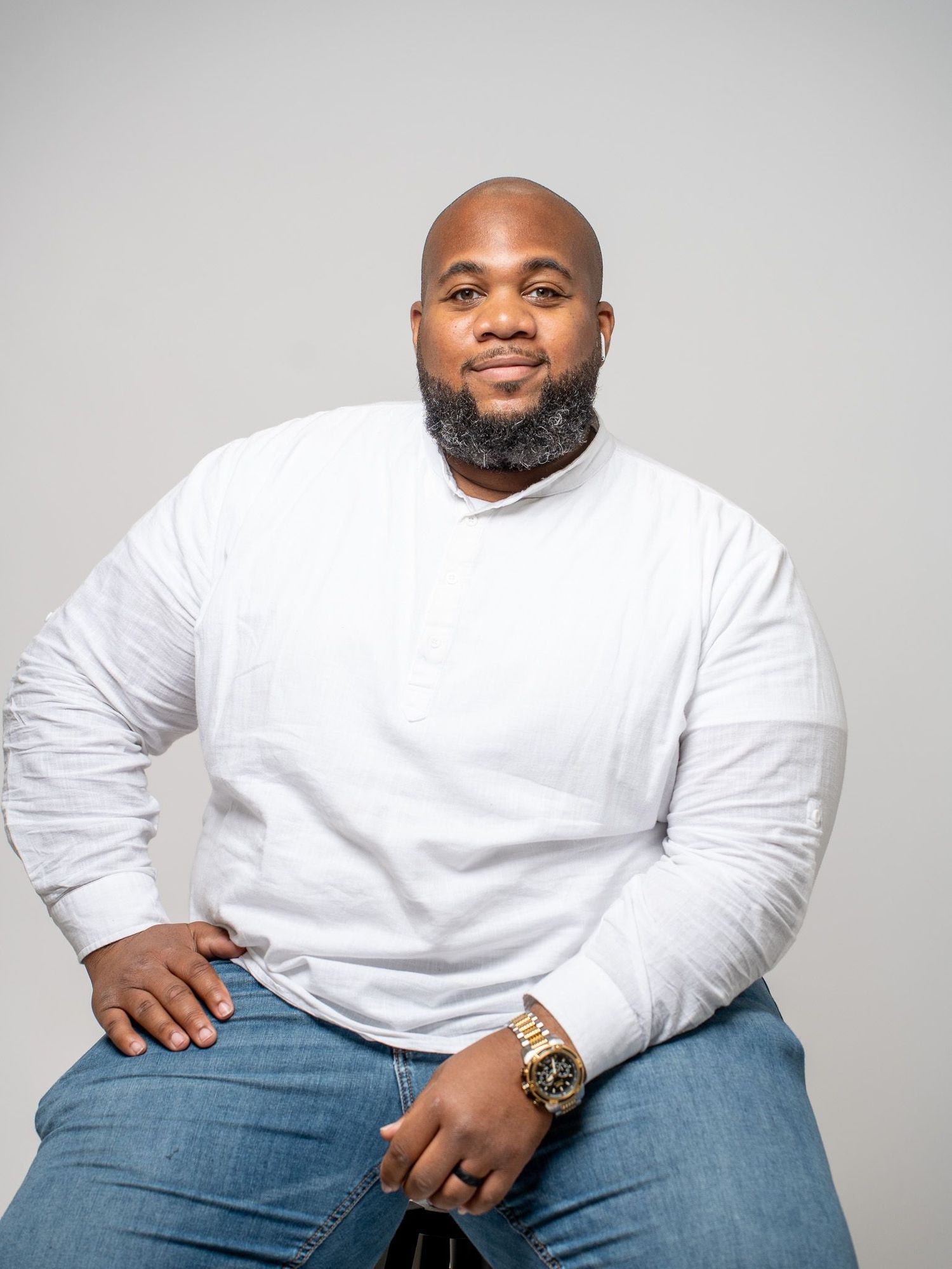 A person wearing a white long-sleeve shirt and jeans sits posed against a gray background while wearing a gold watch.