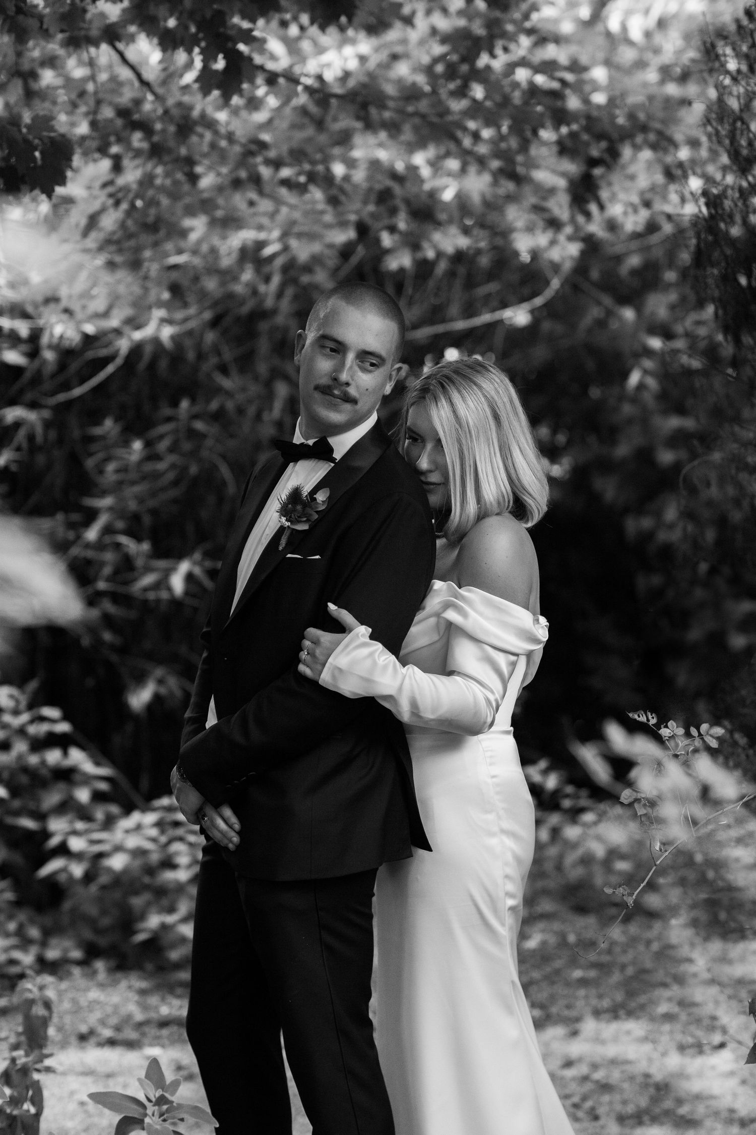 Black and white photo of a couple in wedding attire embracing outdoors with trees in the background.