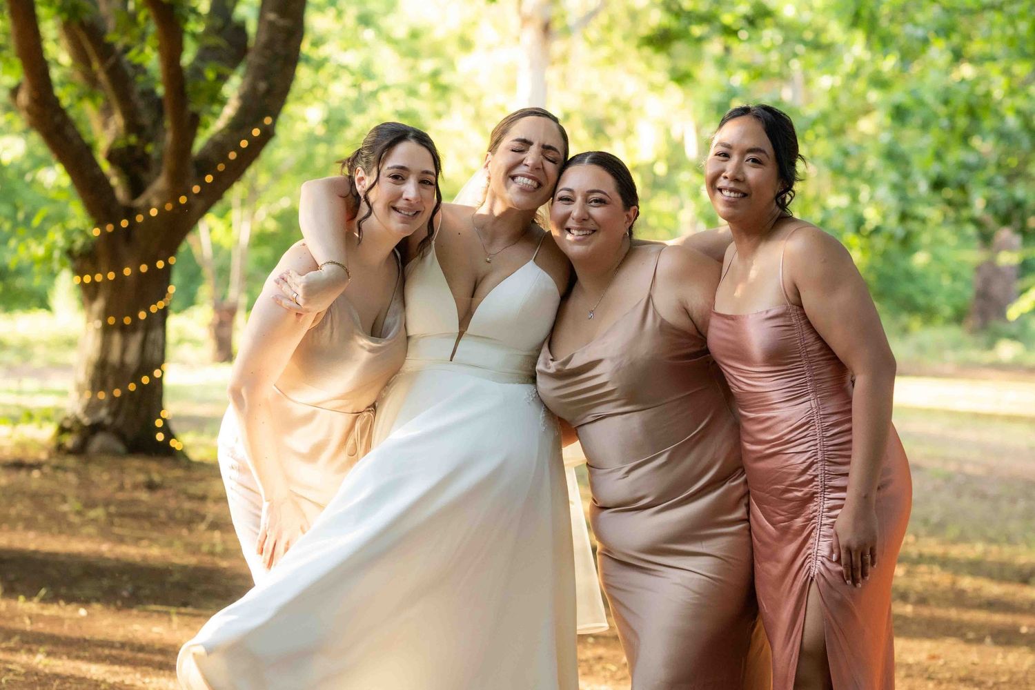 A group of bridesmaids in blush and champagne dresses laugh together under trees at an outdoor wedding celebration.