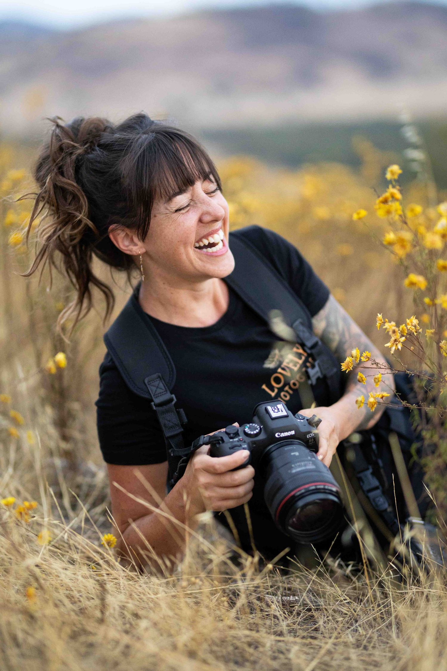 A photographer lies in a field of yellow wildflowers while taking pictures with a professional camera on a sunny day.