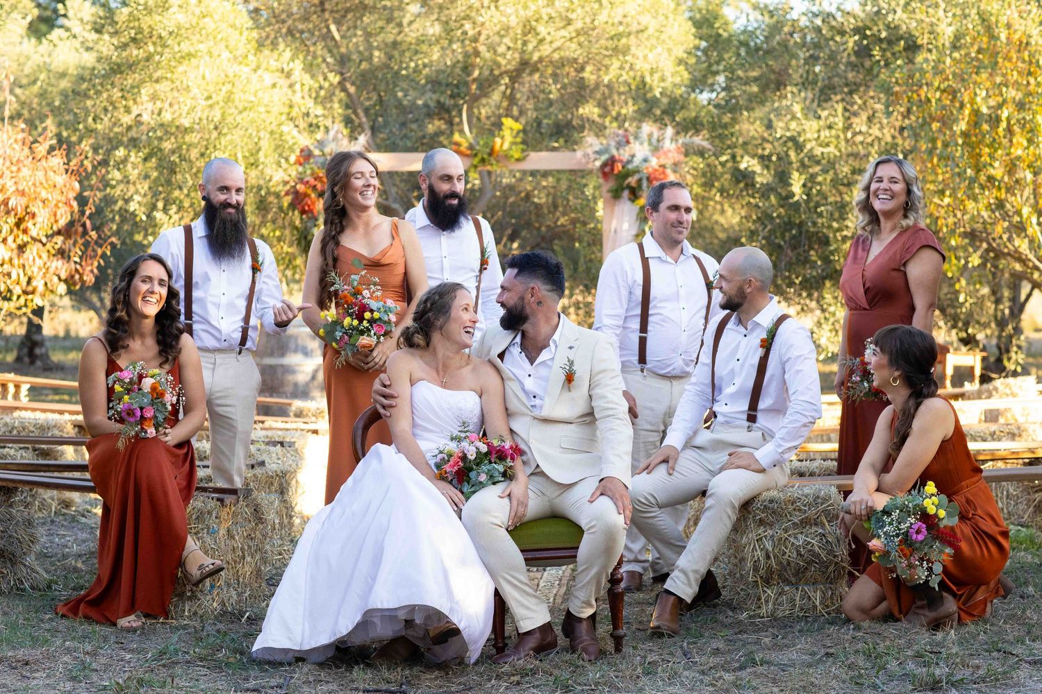 A wedding party in white and rust-colored attire poses together on hay bales during an outdoor autumn celebration.