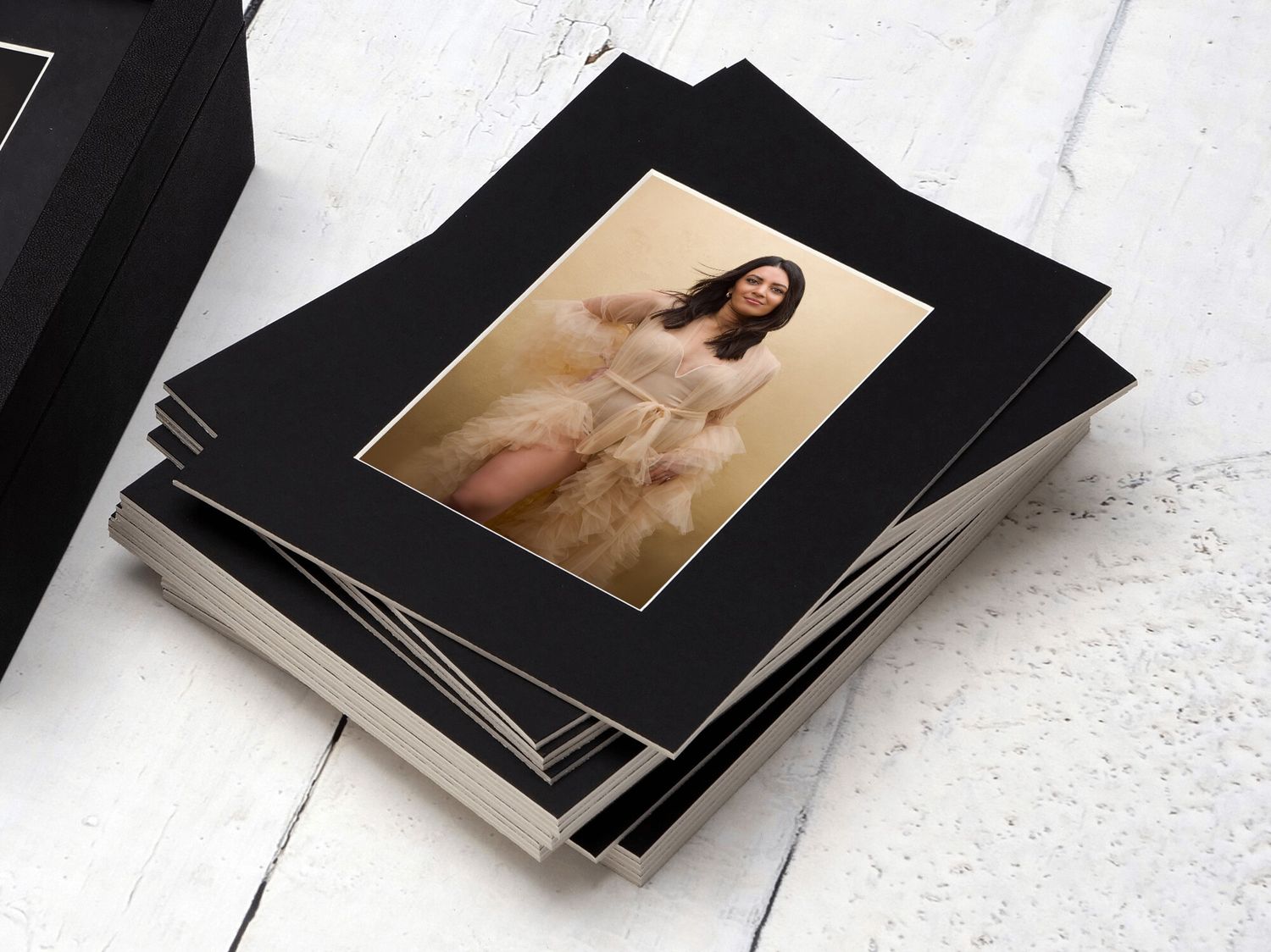 Stack of black matted photo prints featuring a woman in a white dress, displayed on a white wooden surface.