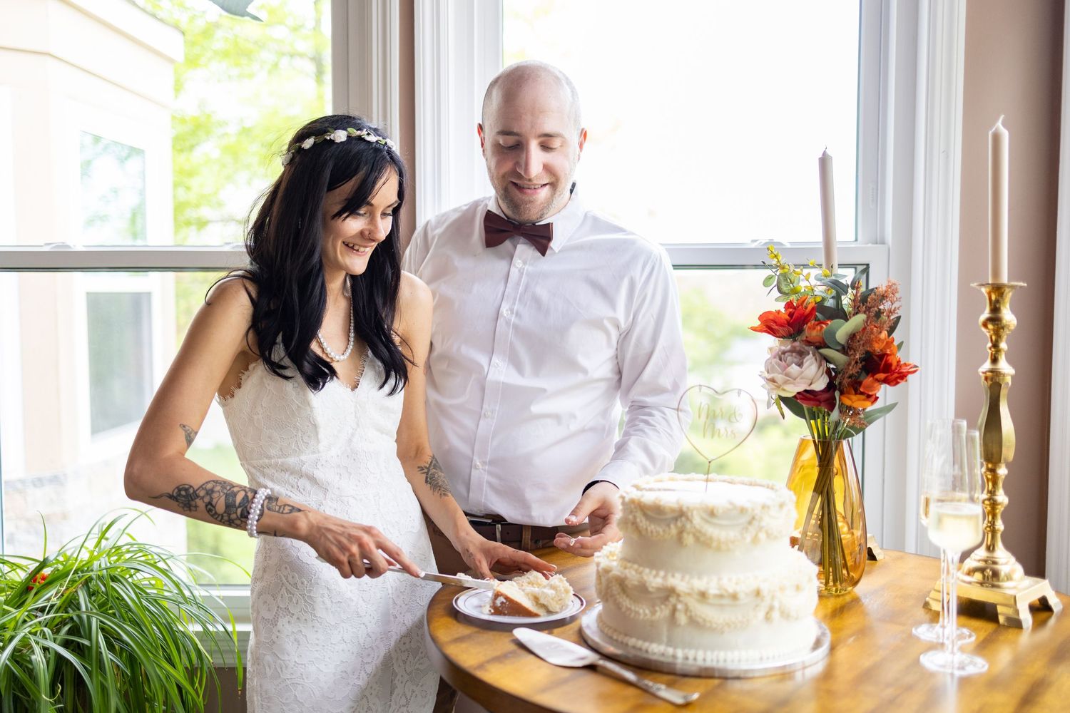 Smiling bride and groom cutting elegant two-tier white wedding cake by a sunny window.
