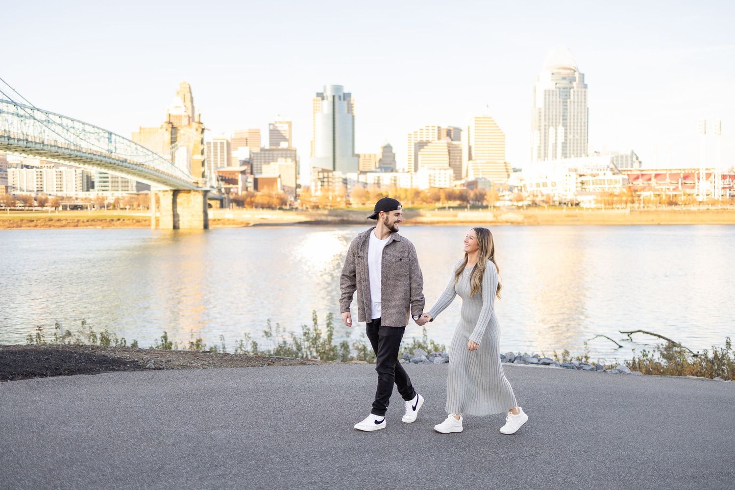 Couple strolling hand in hand along a scenic riverfront path with a city skyline backdrop.