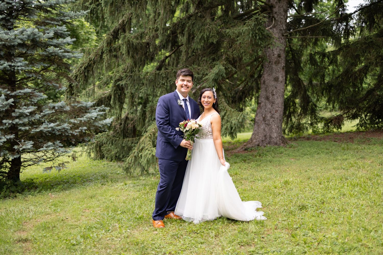 Bride and groom pose together in lush green park setting, bride holding bouquet.