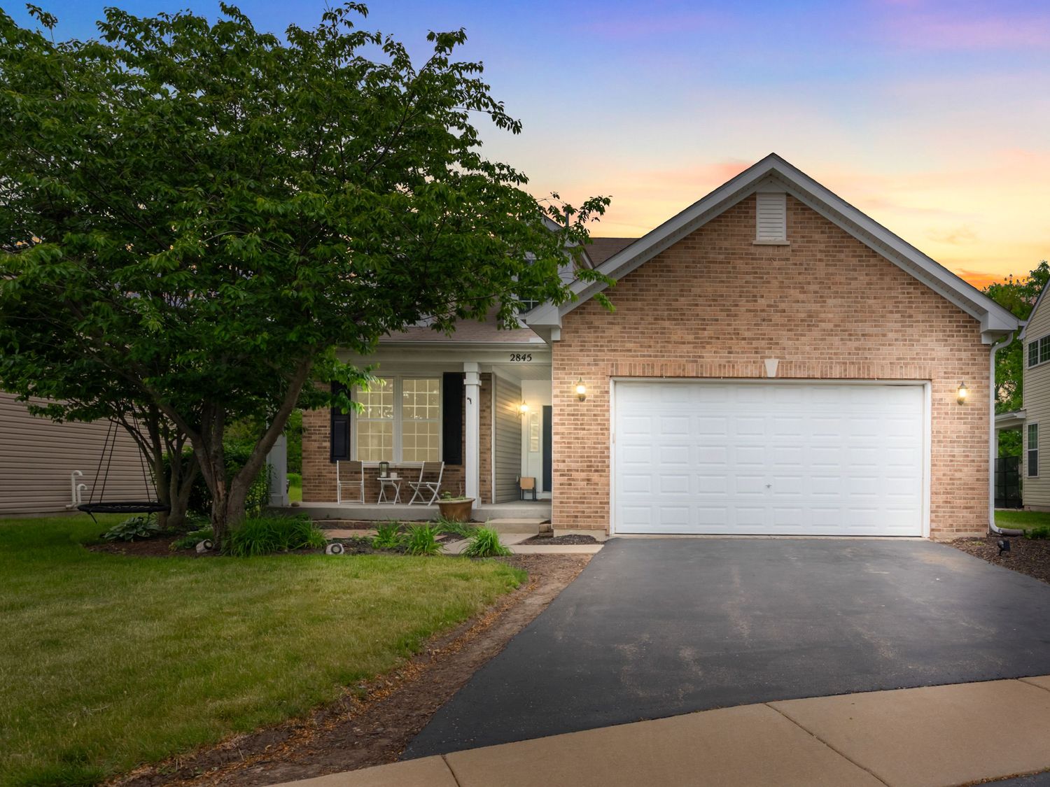 Single-story brick ranch home with white garage door and lush green tree in front yard at sunset.