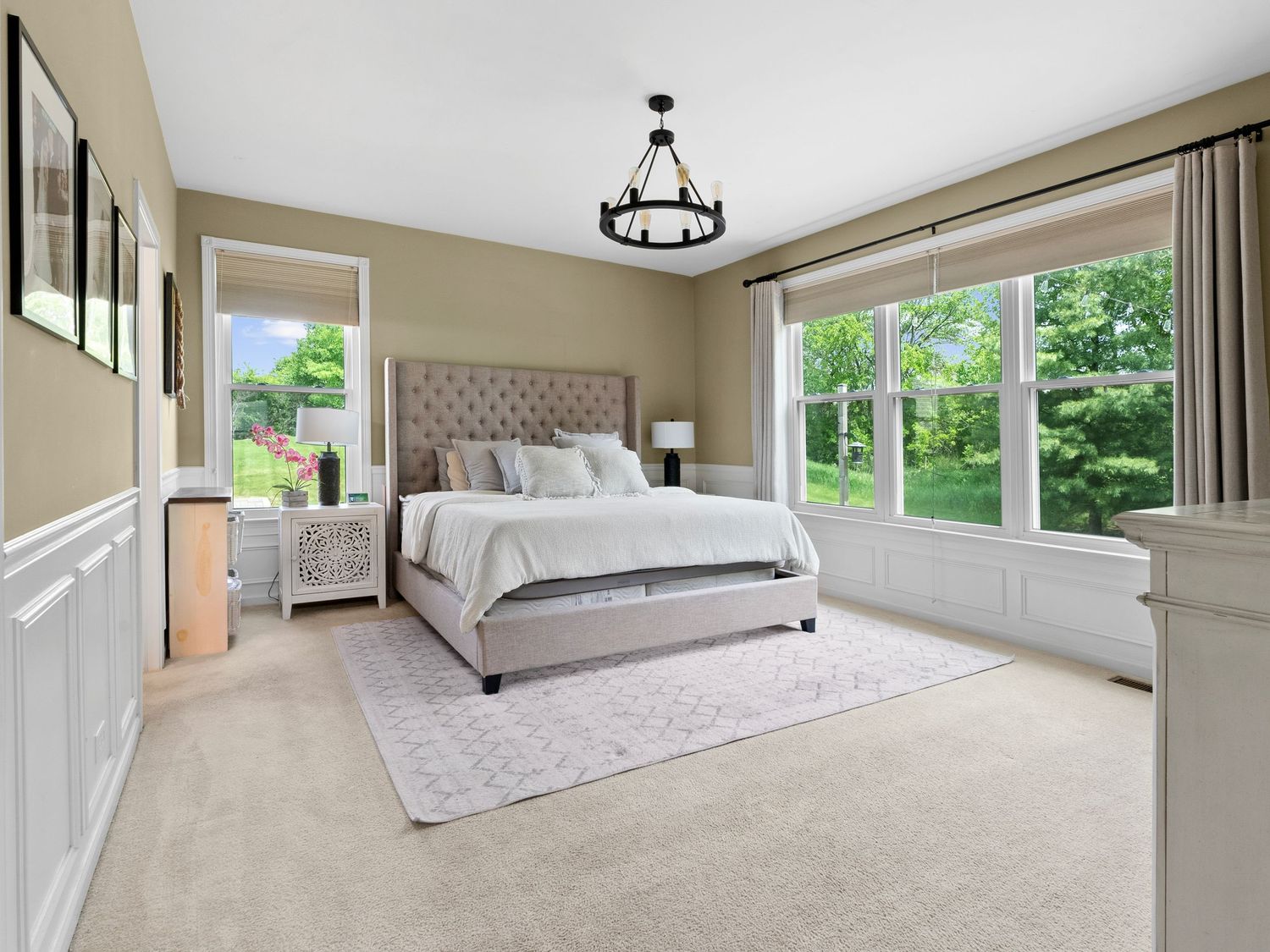 Elegant master bedroom with tufted headboard, beige walls, large windows overlooking greenery, and plush carpeting.