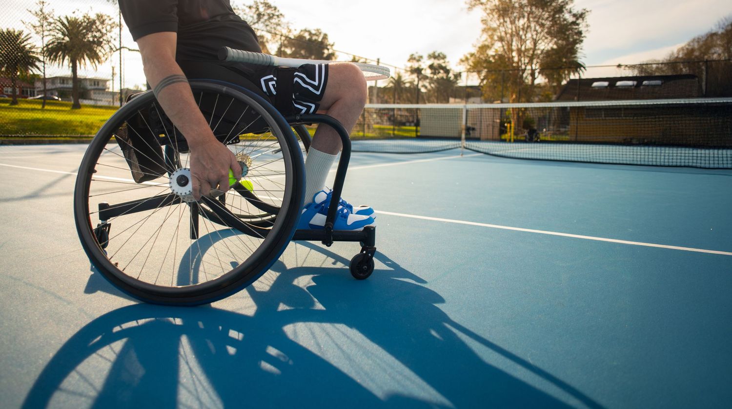 Wheelchair athlete on blue tennis court at sunset casting long shadows during adaptive sports practice.
