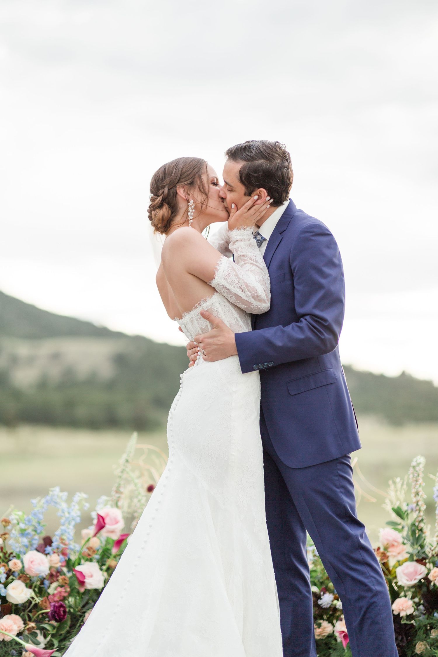 Newlyweds share a romantic kiss in a field with vibrant pink and red flowers during an outdoor wedding ceremony.