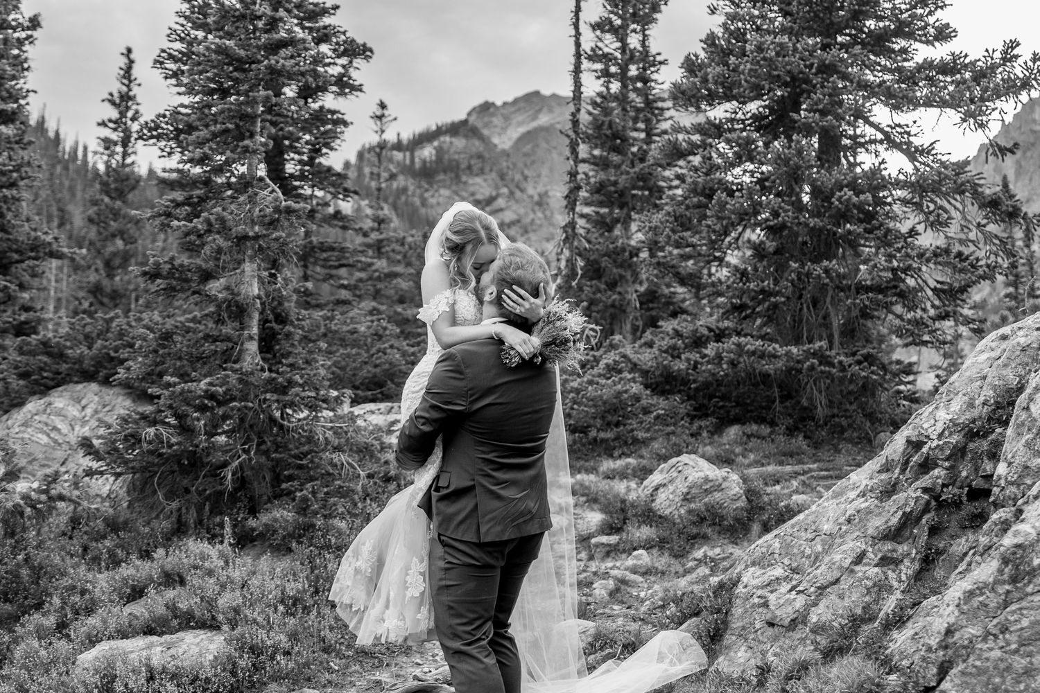 A black and white mountain landscape photo shows evergreen trees and rocky terrain in a rugged wilderness setting.