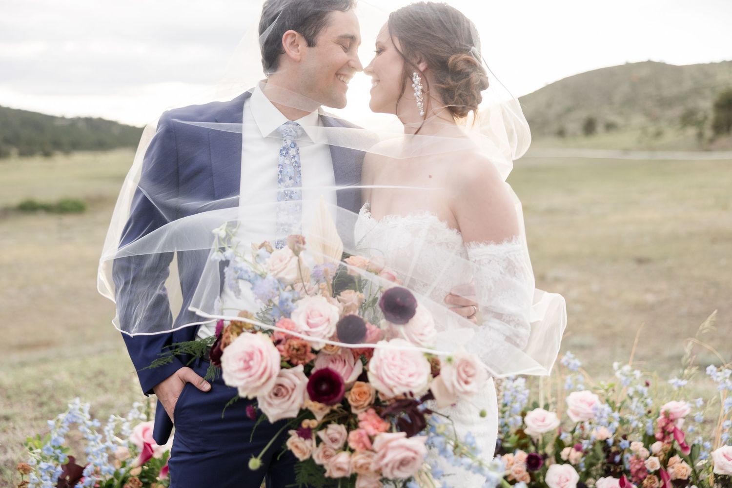 Bride and groom share romantic moment under veil surrounded by lush floral arrangements in an outdoor wedding setting.