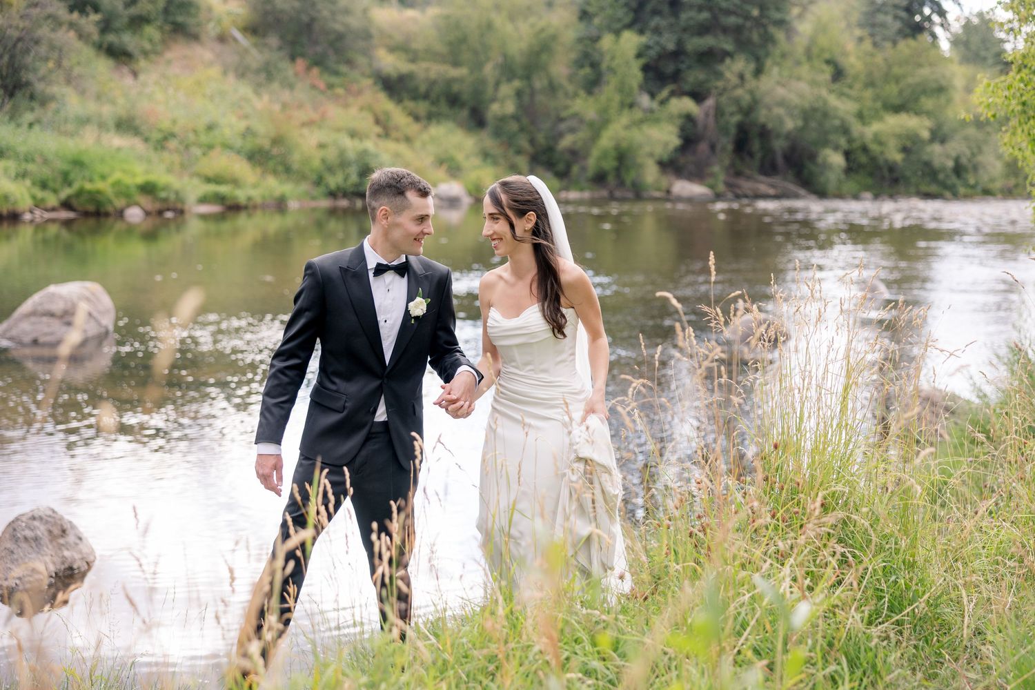 A couple in wedding attire walks through tall grass beside a tranquil pond on their special day.