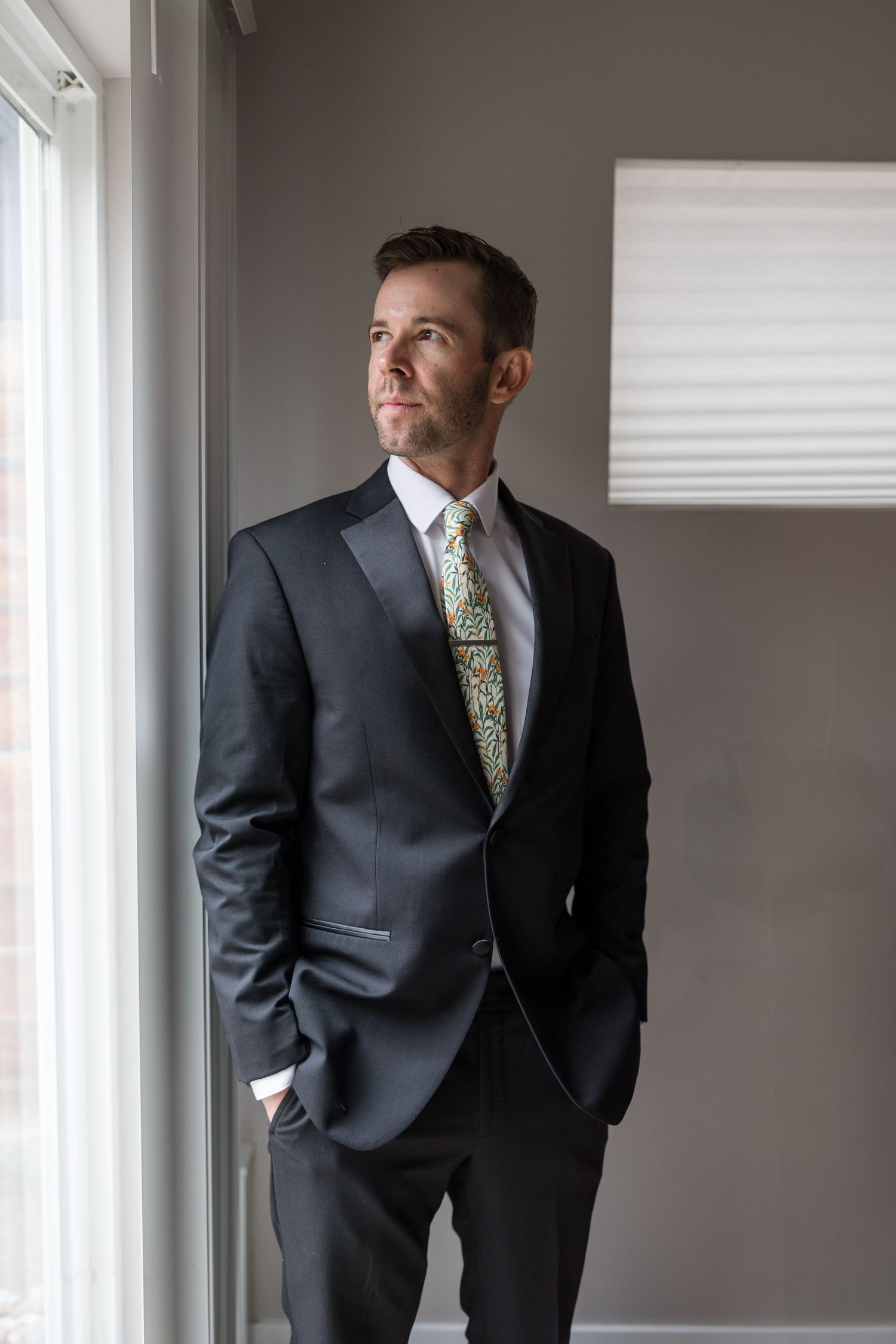Professional in dark suit and patterned tie stands thoughtfully by window in modern office setting.