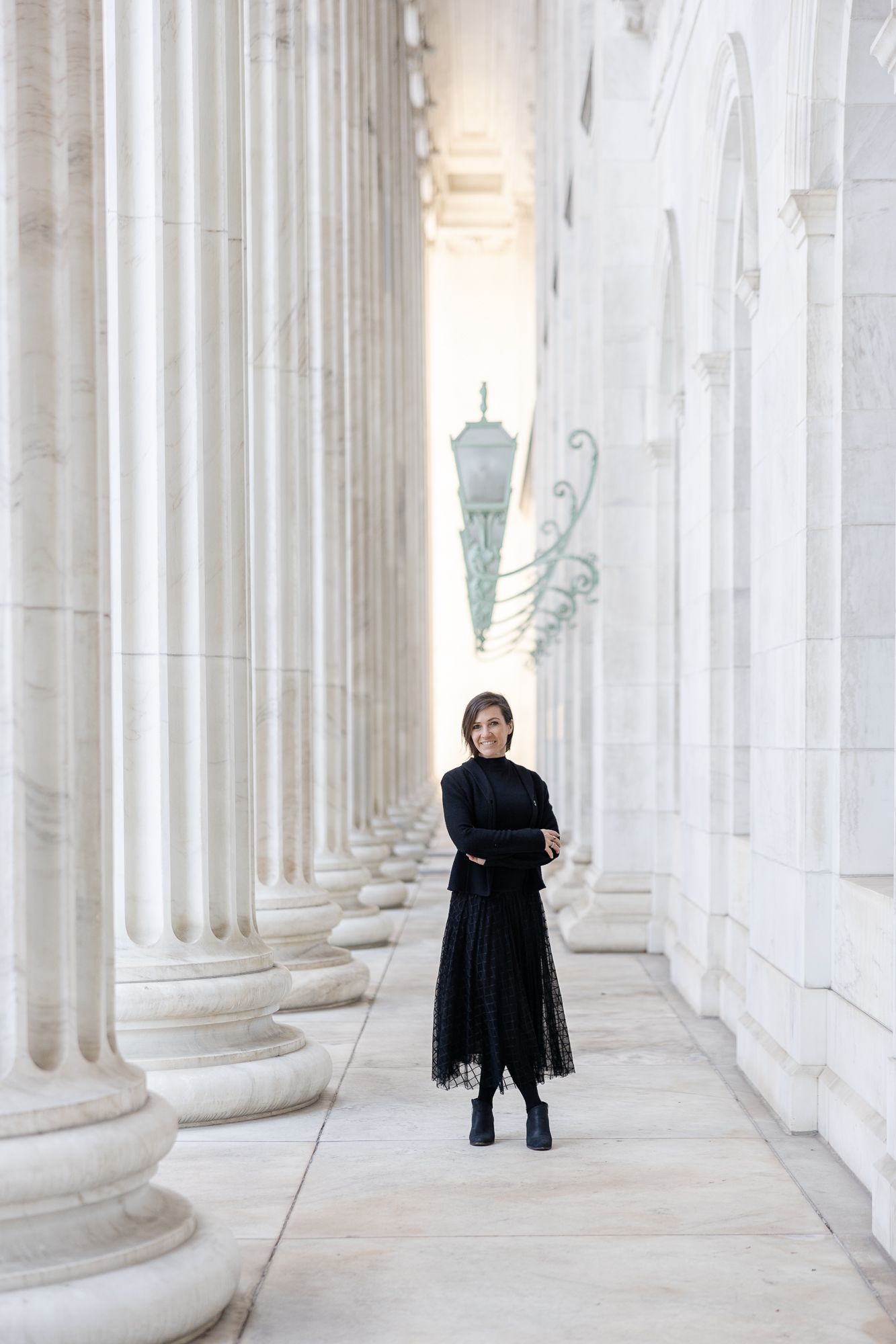 A person in black clothing stands between white marble columns in a classical architectural corridor.
