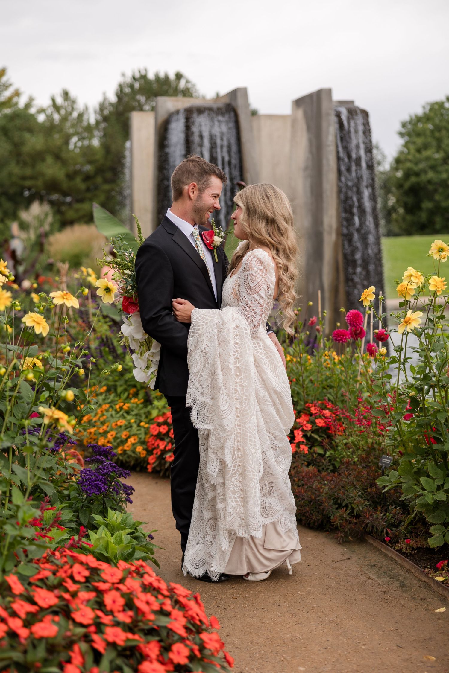 A couple shares a romantic moment in a garden path surrounded by vibrant summer flowers and stone pillars.
