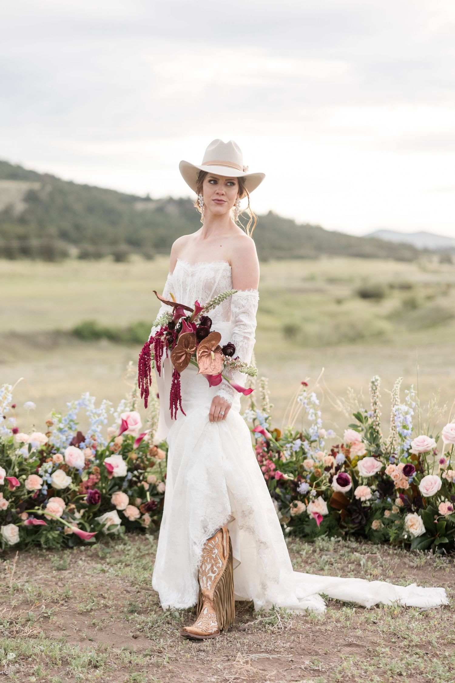 Rustic western bridal portraits in a field with pink and burgundy floral arrangements and cowboy boots.