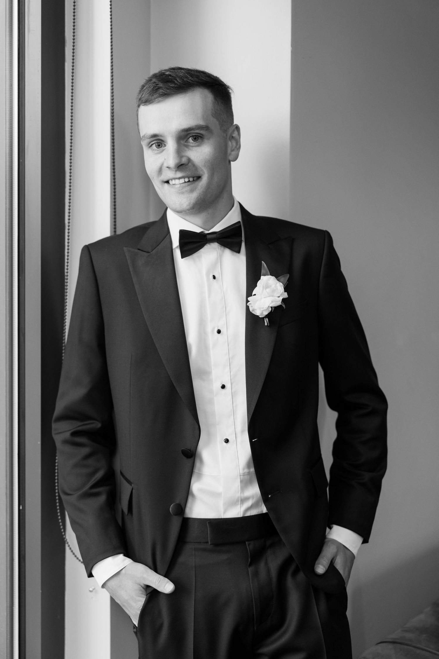 A groom in a classic black tuxedo and white boutonniere poses against a white backdrop in a formal portrait.