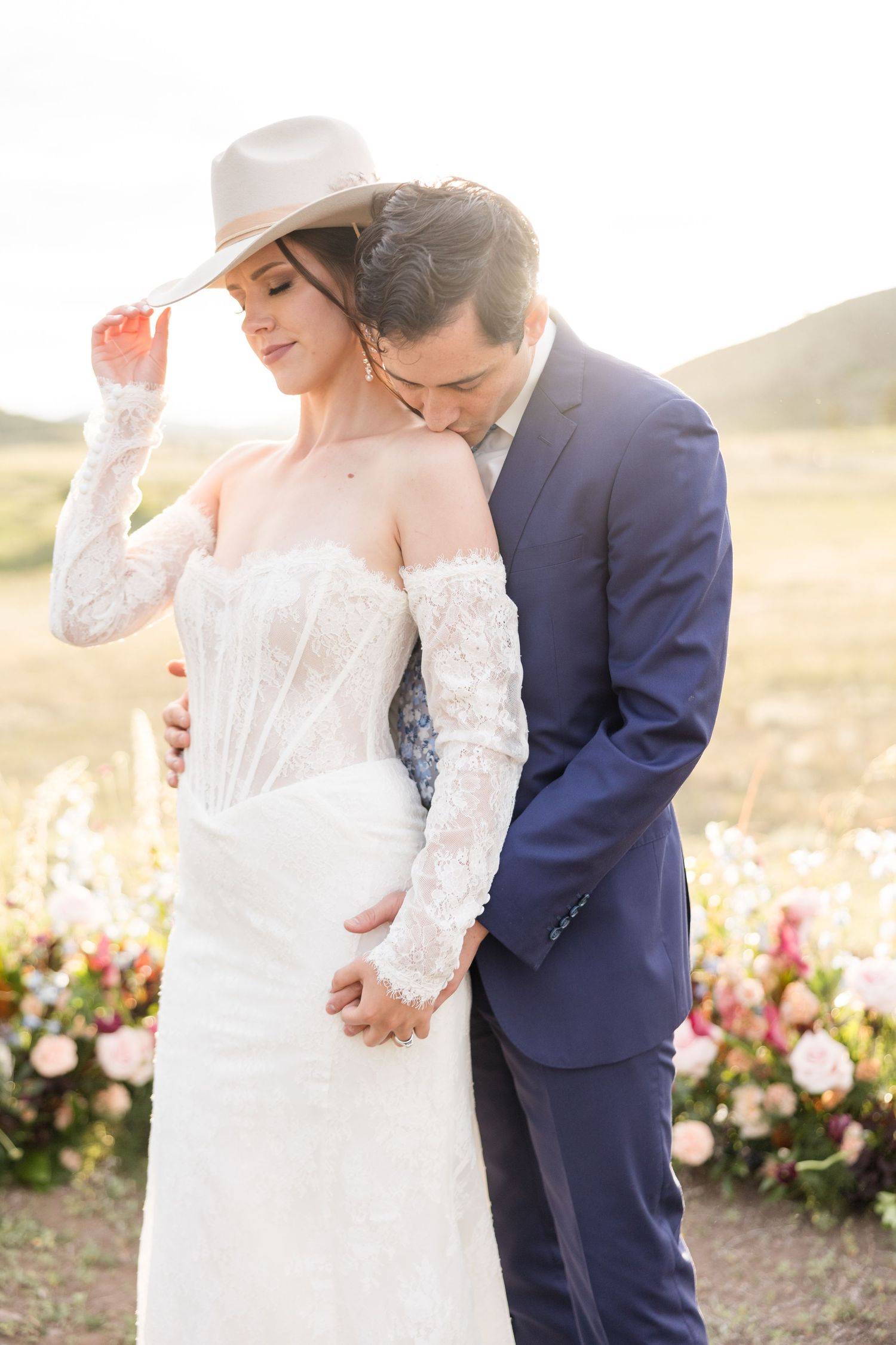 Romantic wedding couple embraces in field of flowers at sunset with bride wearing lace gown and white hat.