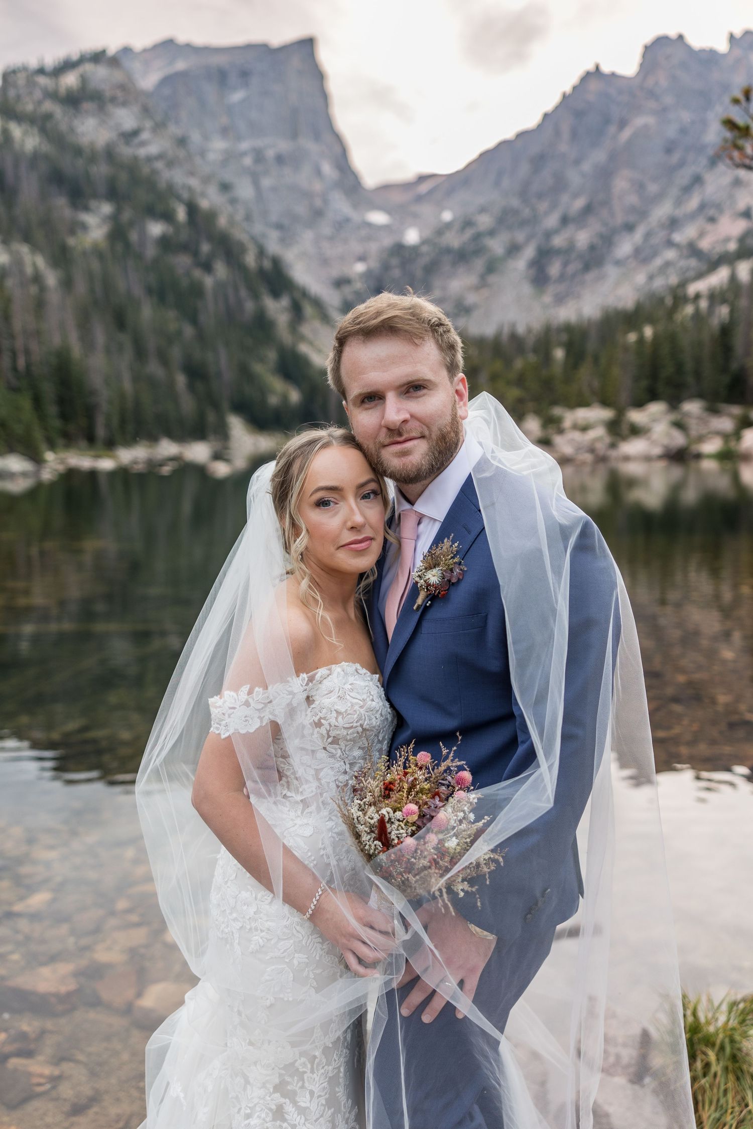 A romantic wedding photoshoot by a serene mountain lake with dramatic rocky peaks in the background.