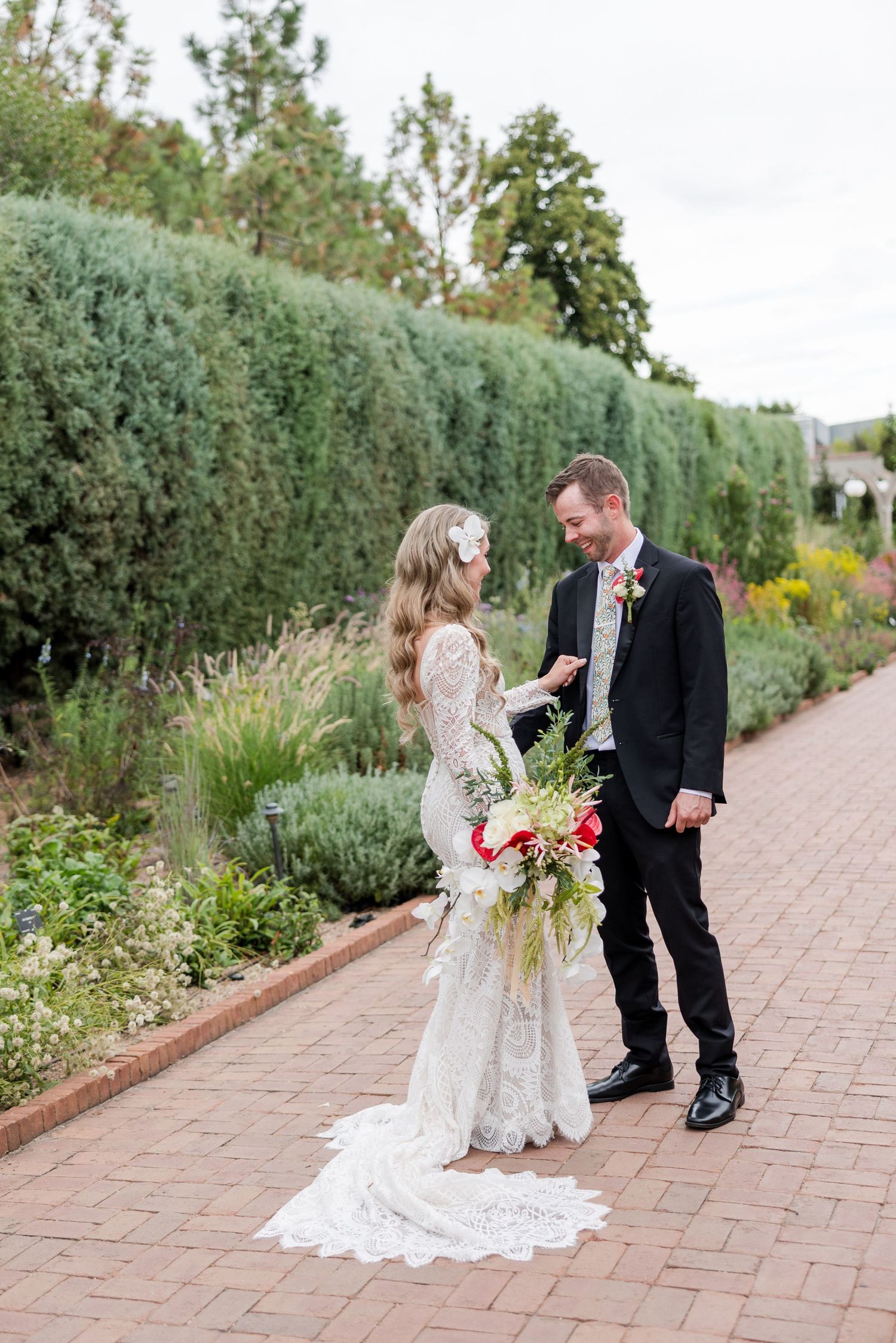 Couple in wedding attire share a romantic moment on a brick path beside lush garden hedges and flowers.
