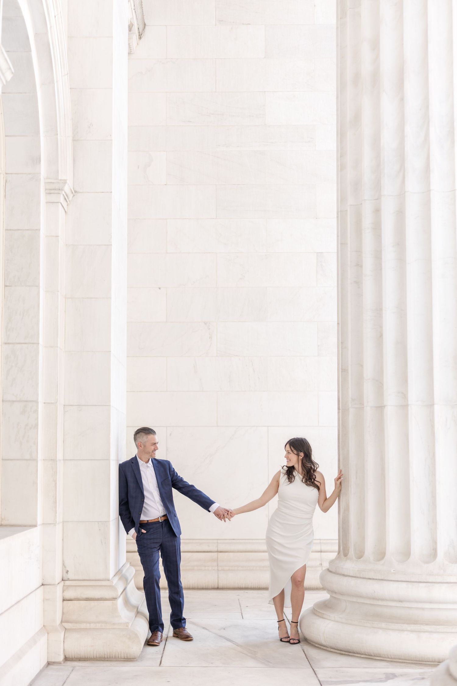A couple in wedding attire playfully poses between white marble columns at a grand architectural venue.