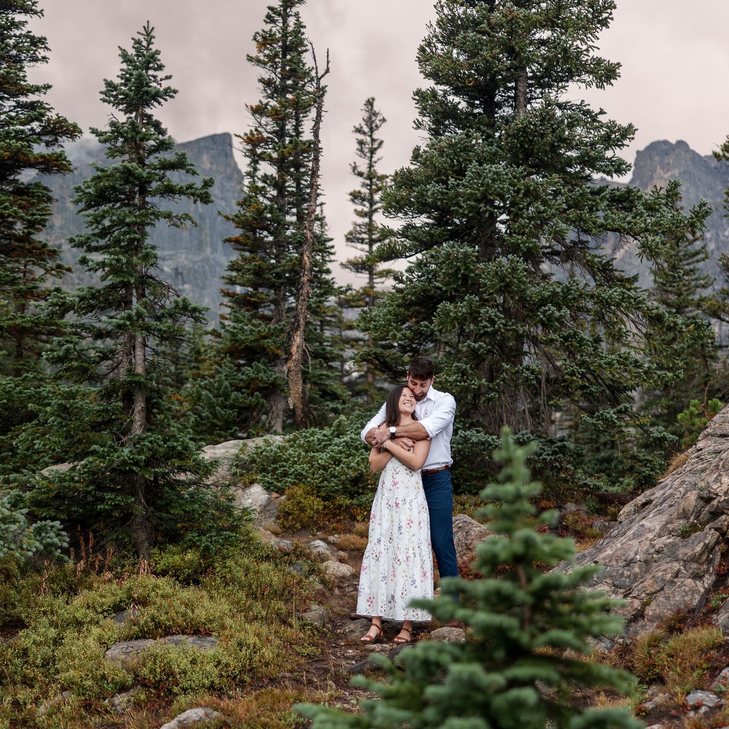 A couple shares an intimate moment among evergreen trees with mountain peaks in the distance on a cloudy day.
