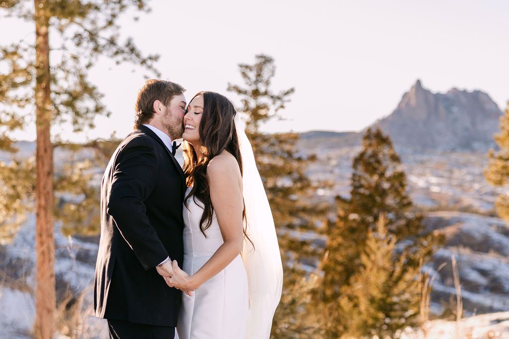 Bride and groom share a romantic kiss outdoors with rocky mountain landscape and pine trees in winter backdrop.