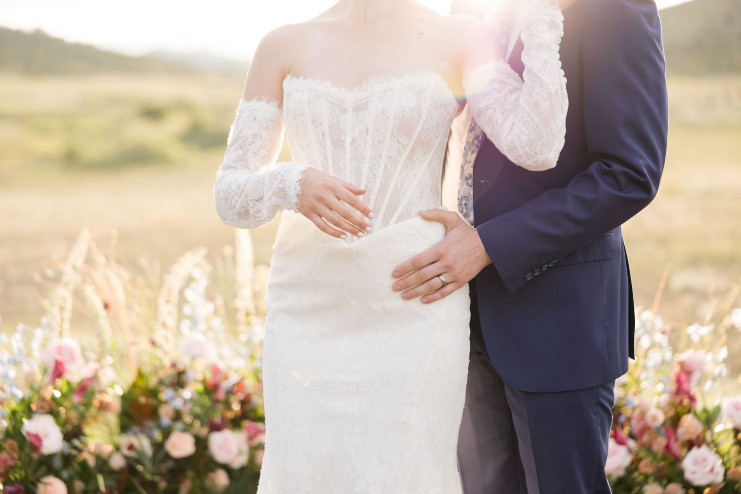 Couple embraces at sunset by floral wedding arch in field with bride wearing lace gown and groom in navy suit.