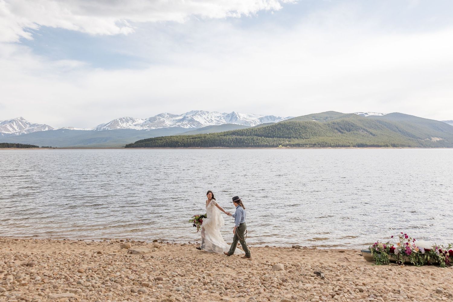 Snow-capped mountains rise above a tranquil lake with a rocky shoreline in a scenic mountain landscape.