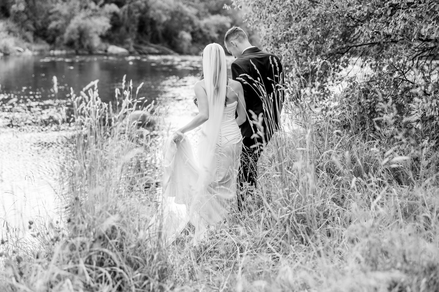 Black and white romantic photograph of a couple standing in tall grass by a serene lake.