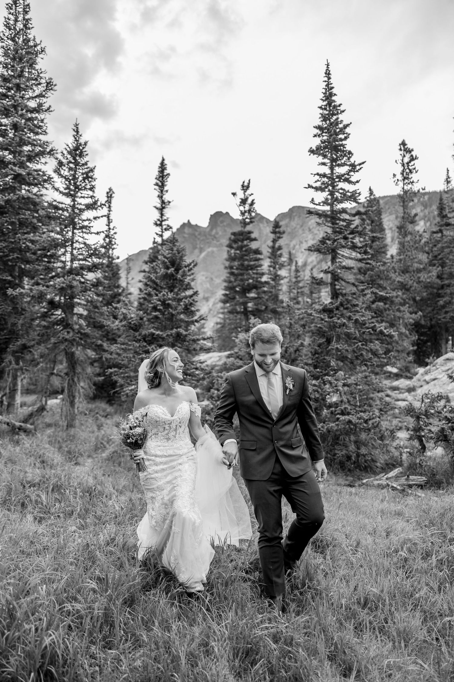 A couple walks together through a mountain meadow with evergreen trees and peaks in the background.