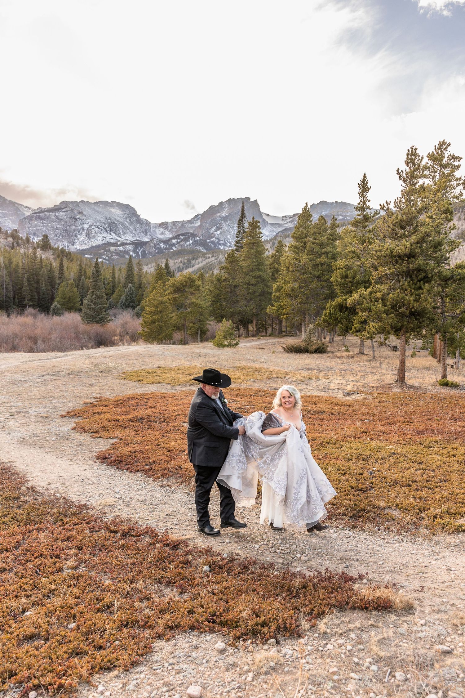 Two figures in formal attire share a romantic moment on a mountain path surrounded by autumn foliage and snow-capped peaks.