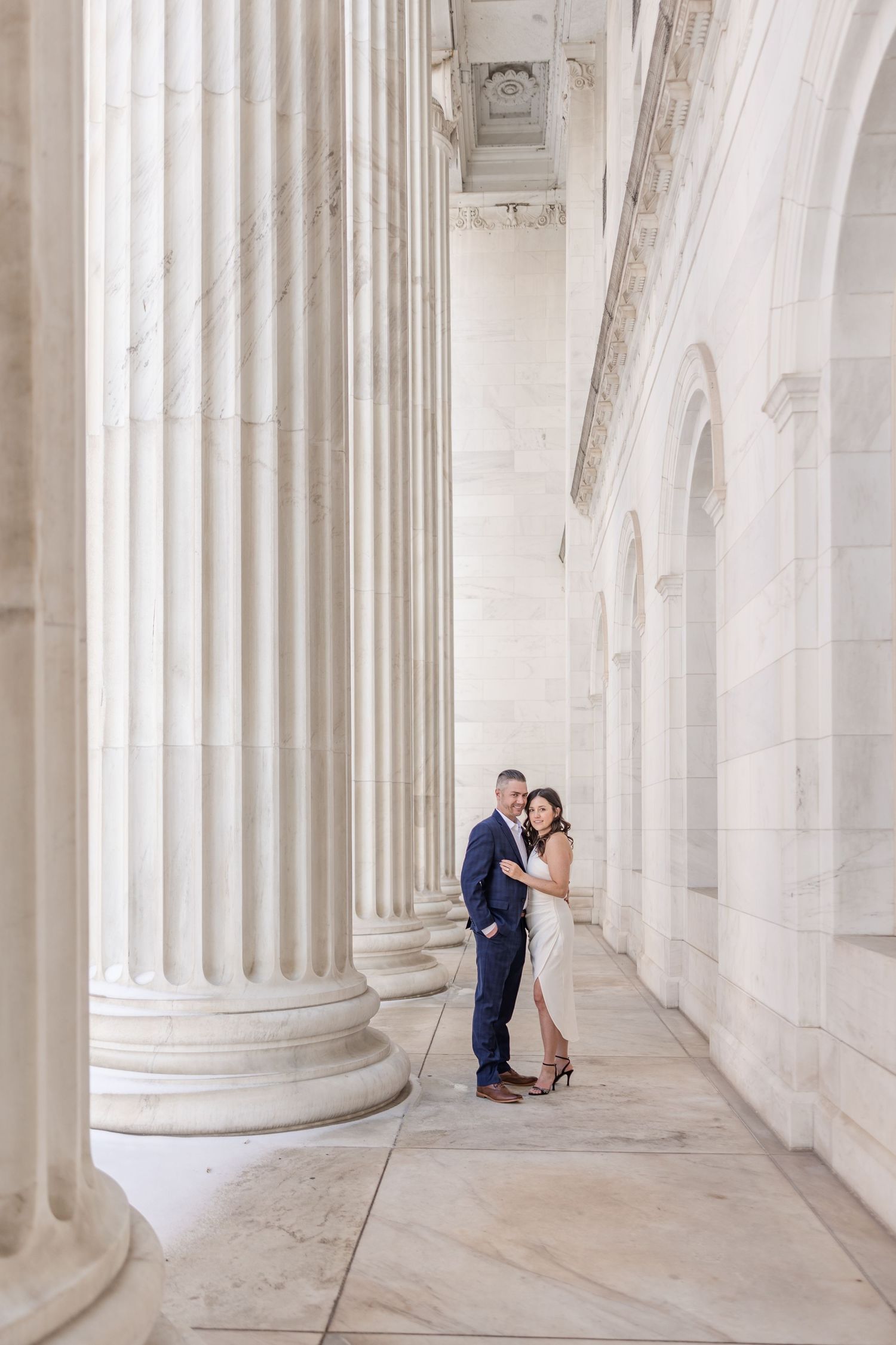 A couple poses romantically between white marble columns in an elegant neoclassical architectural setting.