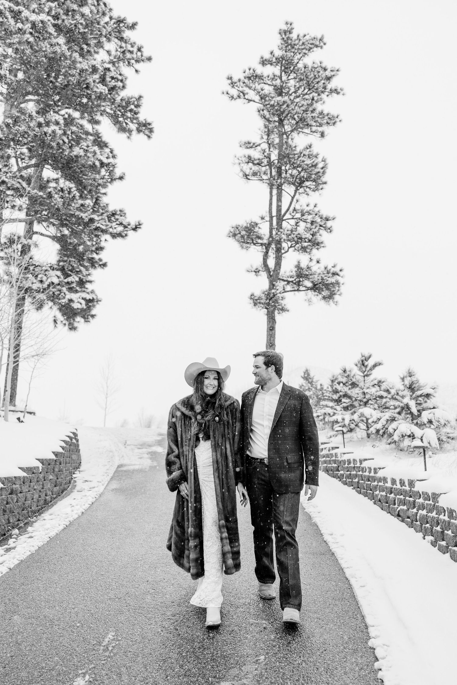 Black and white photo of a couple walking together on a snowy path surrounded by snow-covered pine trees.