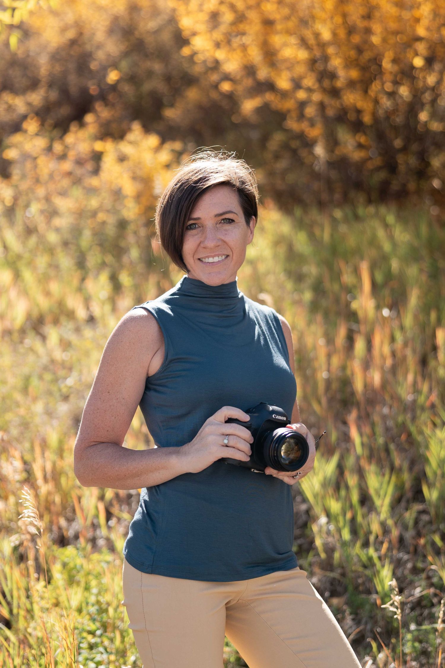 A photographer in khaki pants and navy shirt poses with their camera in a golden sunlit field during autumn.