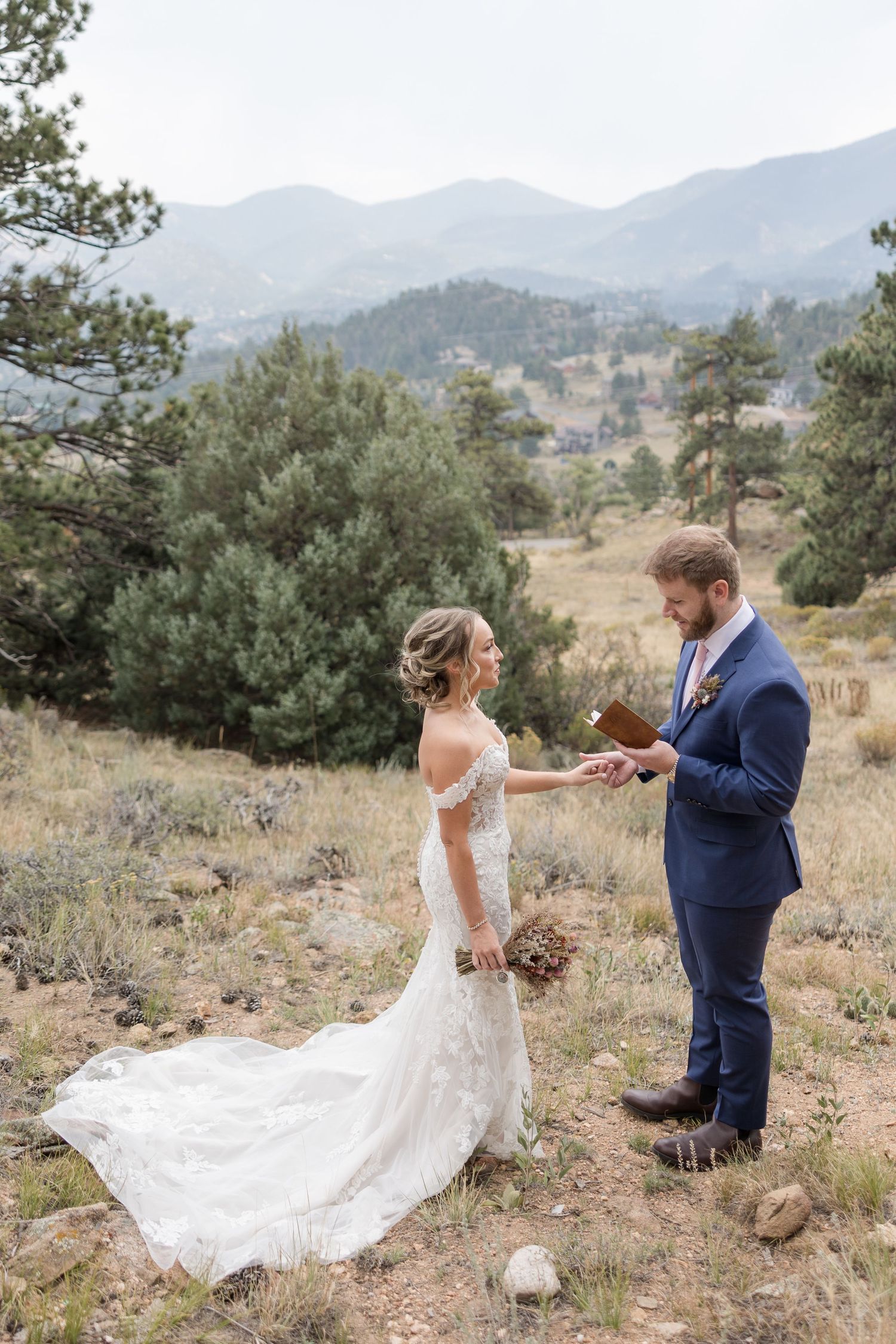 A couple dances together on a mountainside with the bride's white dress flowing in the wind against a scenic landscape.