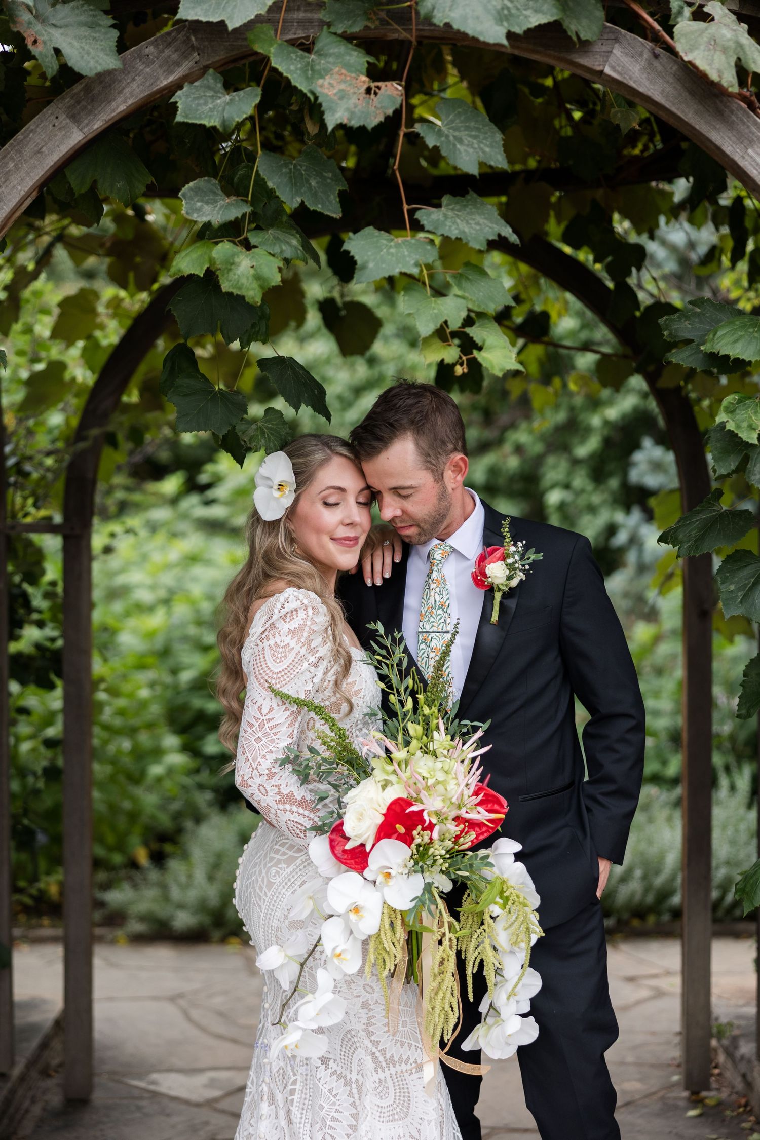 Romantic wedding couple embraces under lush green grape vines in an elegant garden setting.