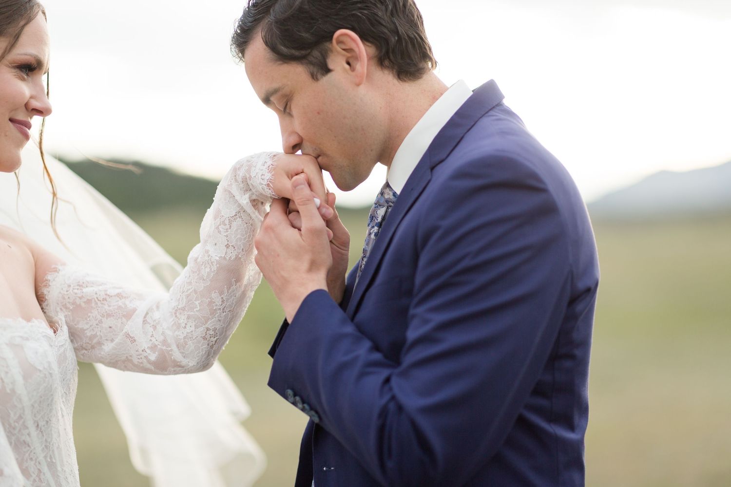 A romantic wedding scene showing a groom gently kissing his bride's hand in a natural setting.