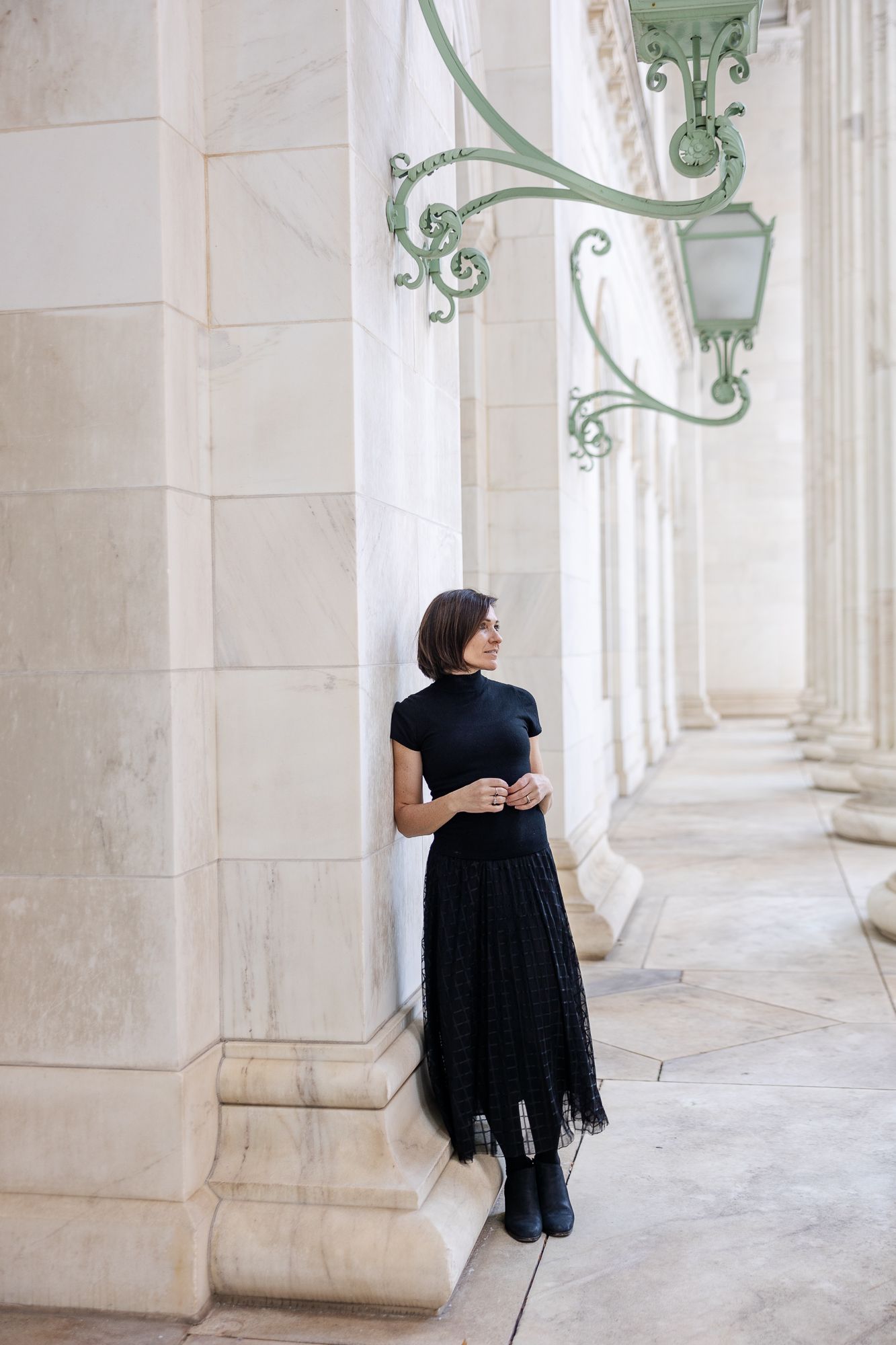 Woman in black dress and boots leaning against marble column of elegant building with ornate green lanterns.