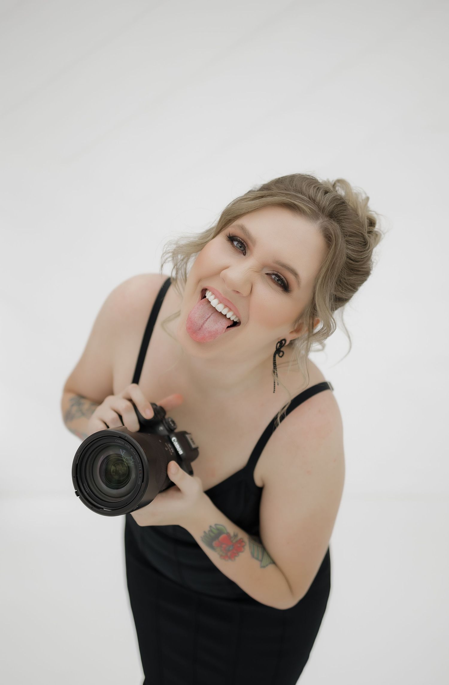 Smiling blonde female photographer holding DSLR camera with tongue out against white studio background.