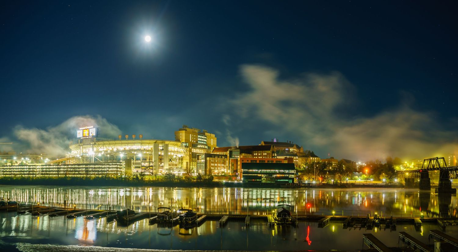 Moonlit cityscape reflecting on calm harbor waters at night with glowing buildings and dramatic cloudy sky.