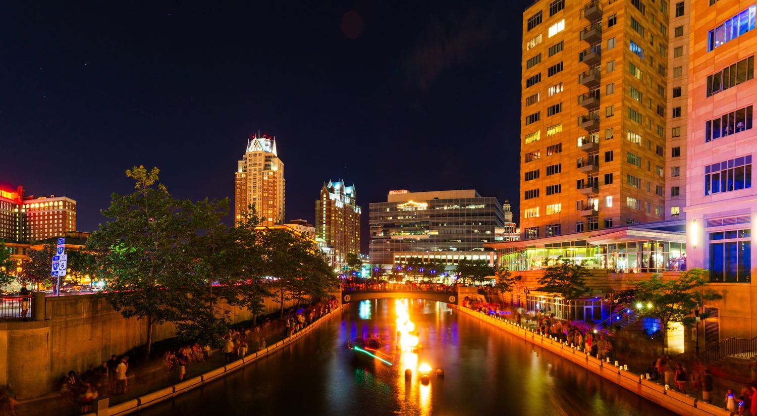 Vibrant city skyline reflecting on a river at night with glowing fire features and illuminated buildings.