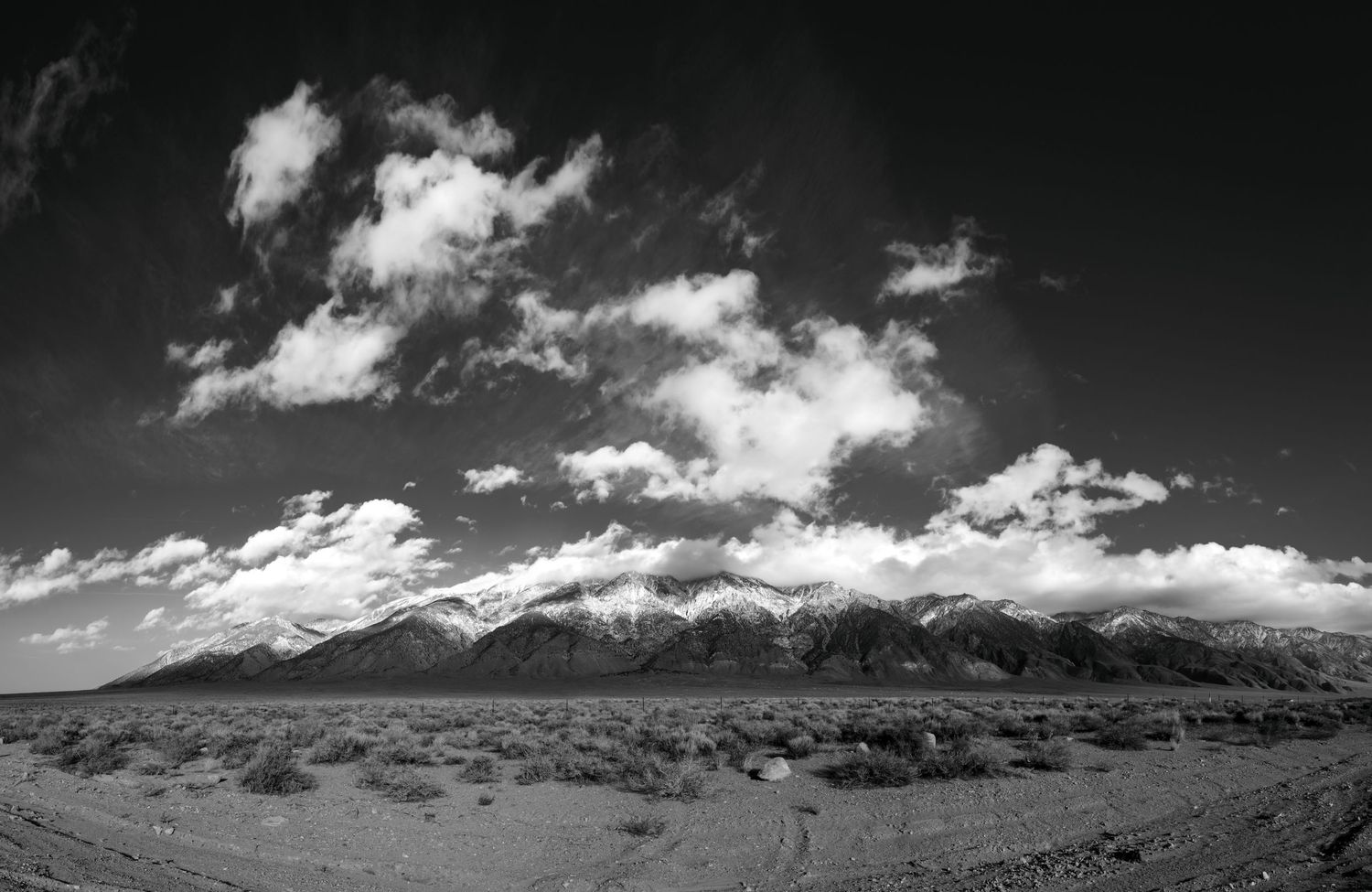 Black and white desert landscape with snow-capped mountains and dramatic cloudy sky.