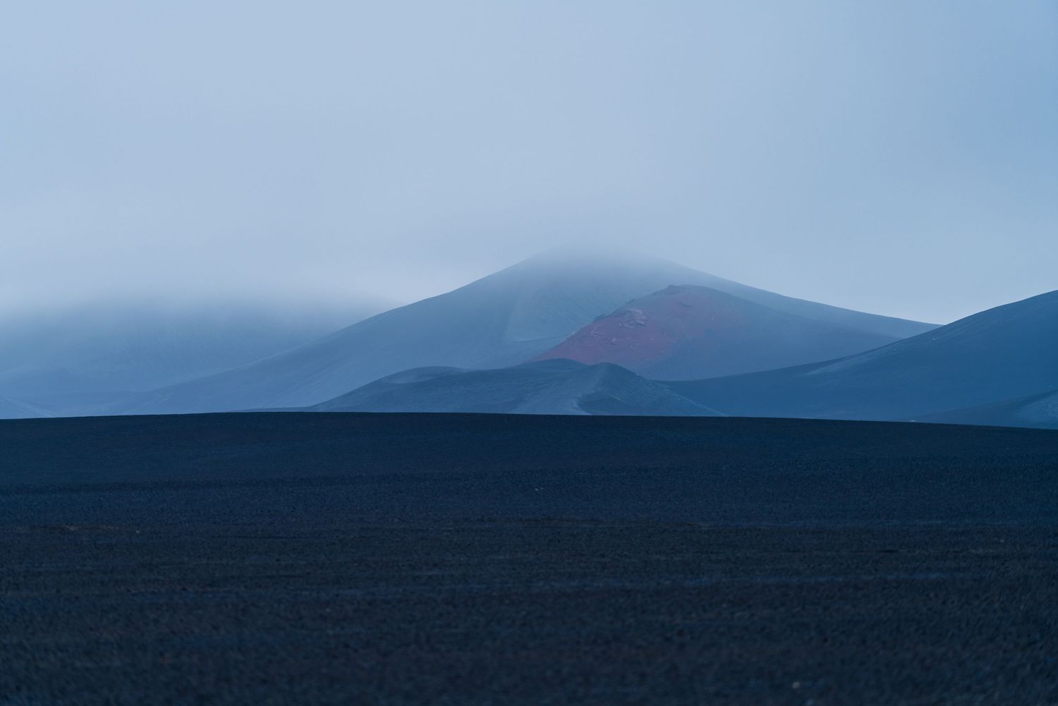 Misty volcanic mountain with red crater rising above dark lava fields under a blue-grey sky in Iceland.
